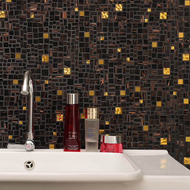Black and gold square mosaic tiles in irregular arrangement on the wall behind cosmetics and a sink