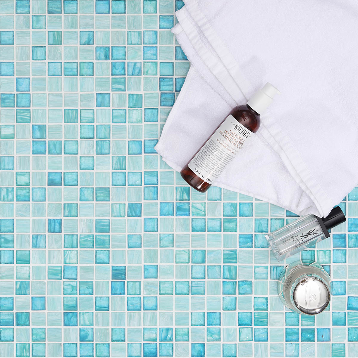 Front view of bright green glass mosaic tiles with a white towel and cosmetics bottle.