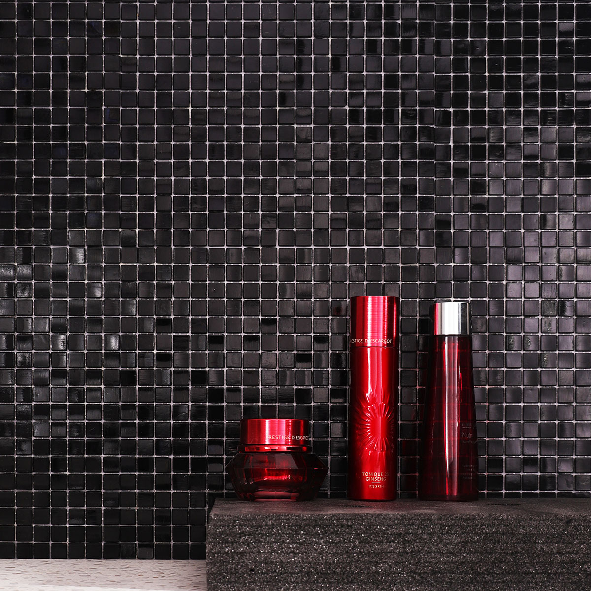 Pure black glass mosaic tiles on the wall with red skincare bottle in front.