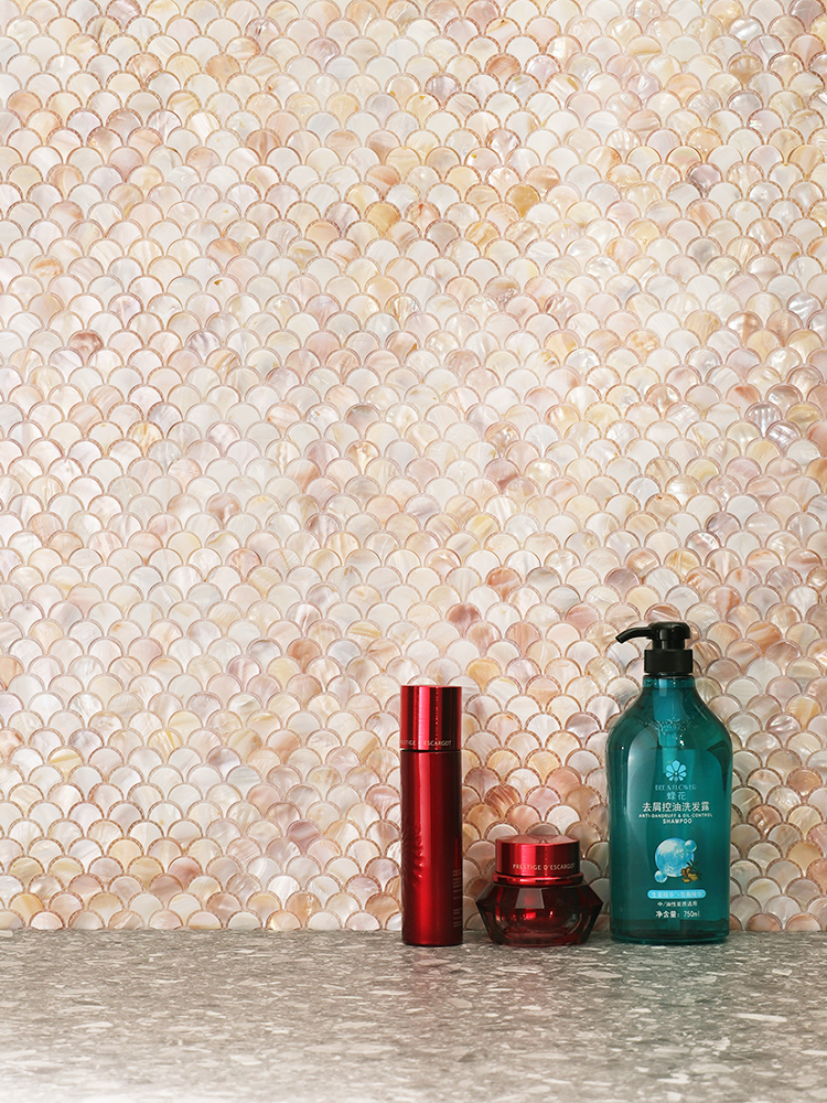 The fish scale shell mosaic adds a luxurious texture to the shower walls, perfectly complemented by neatly placed skincare products and shower gel for a refined bathroom look.