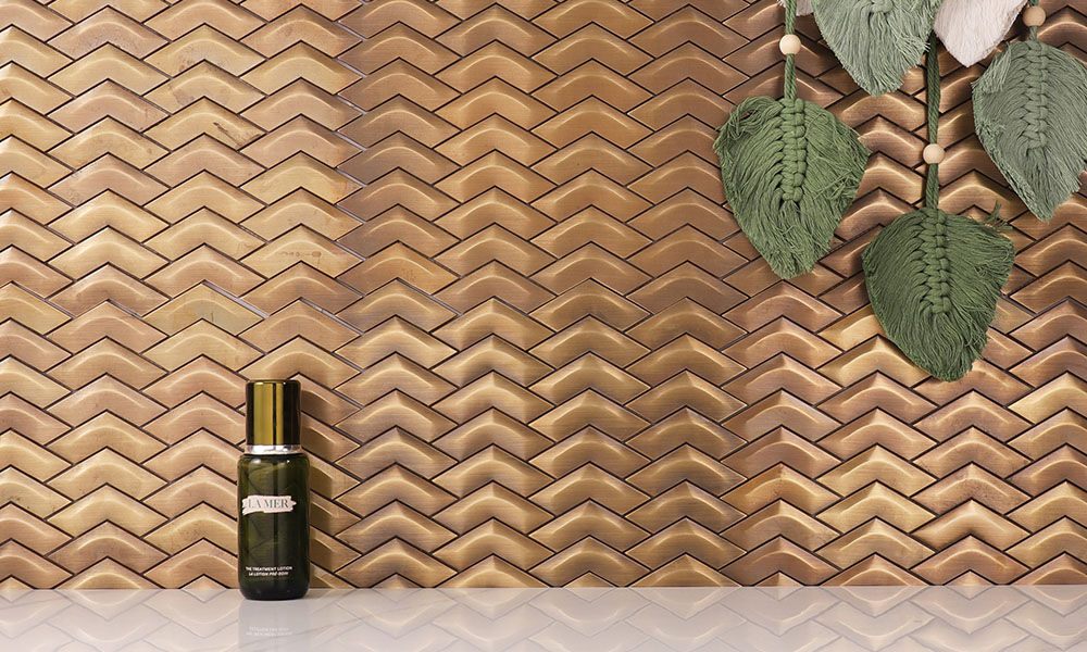 Fish scale copper mosaic tile on the wall with a bottle and woven leaves in front.