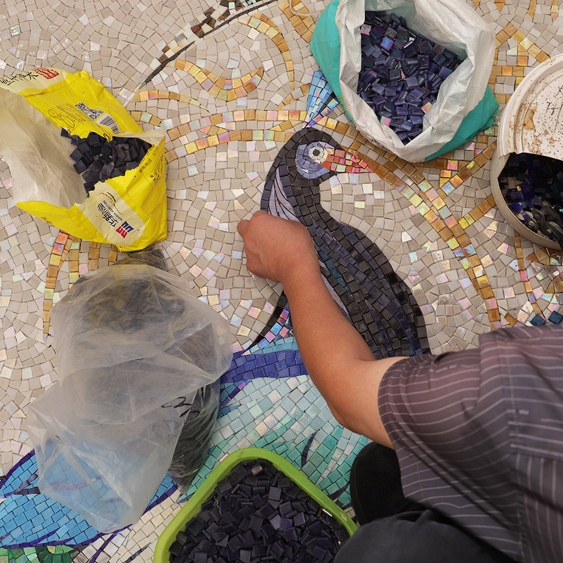 Artisan placing dark glass tiles on a handmade peacock mosaic mural