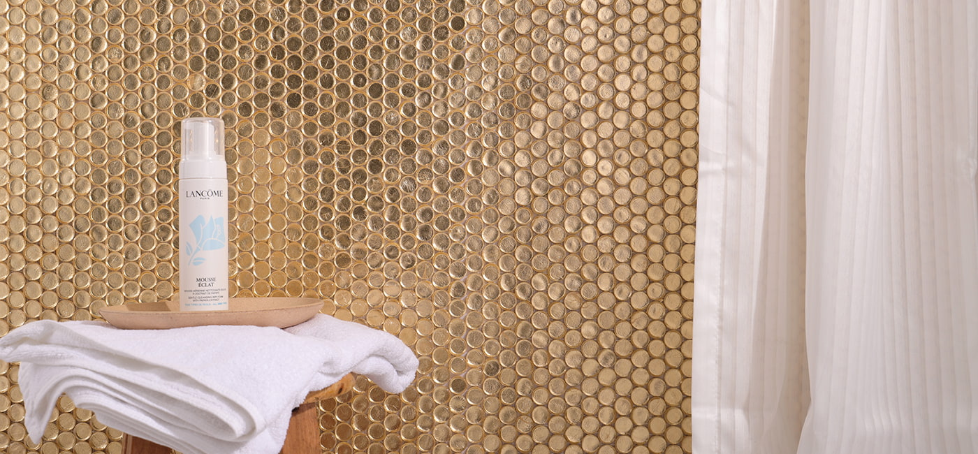 A golden mosaic wall with towels and skincare products on a stool. 