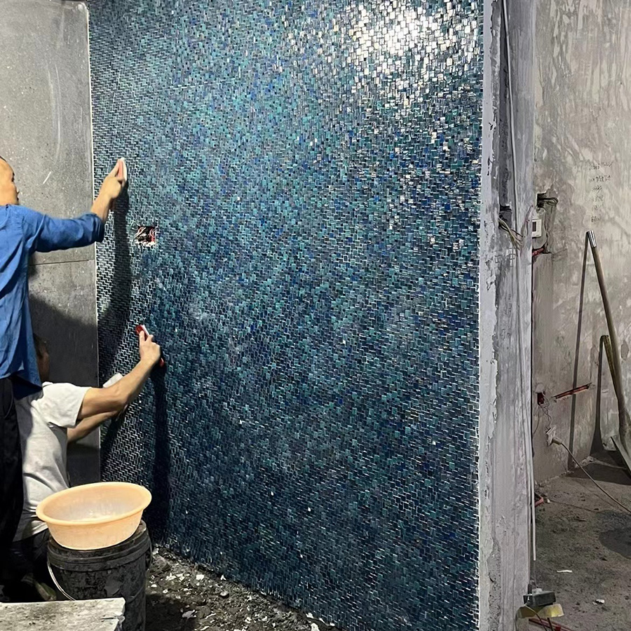 Workers installing blue glass mosaic tile backsplash on interior wall.