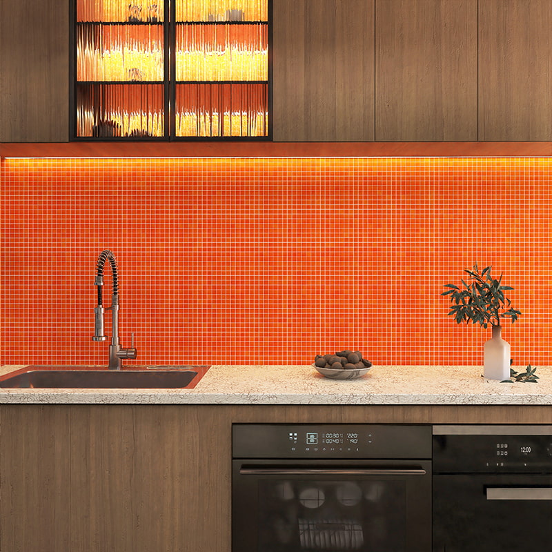 Vibrant orange glass mosaic backsplash illuminated by under-cabinet lighting above marble countertop