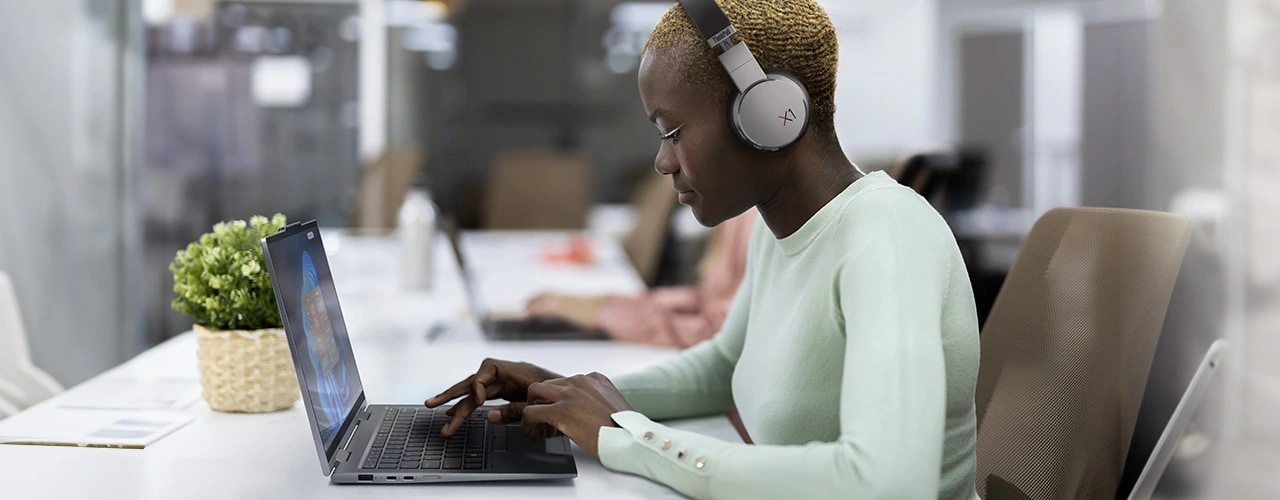 A woman wearing a pair of ThinkPad X1 Active Noise Cancellation Headphones sits at a table typing on the Lenovo ThinkPad X1 2-in-1 convertible laptop, which is in left-side profile view.