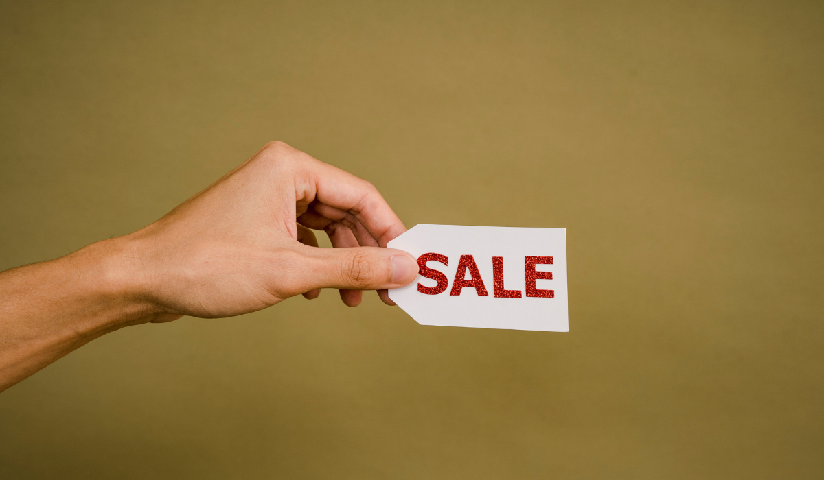 Hand holding a sale tag with red glitter letters on a plain brown background