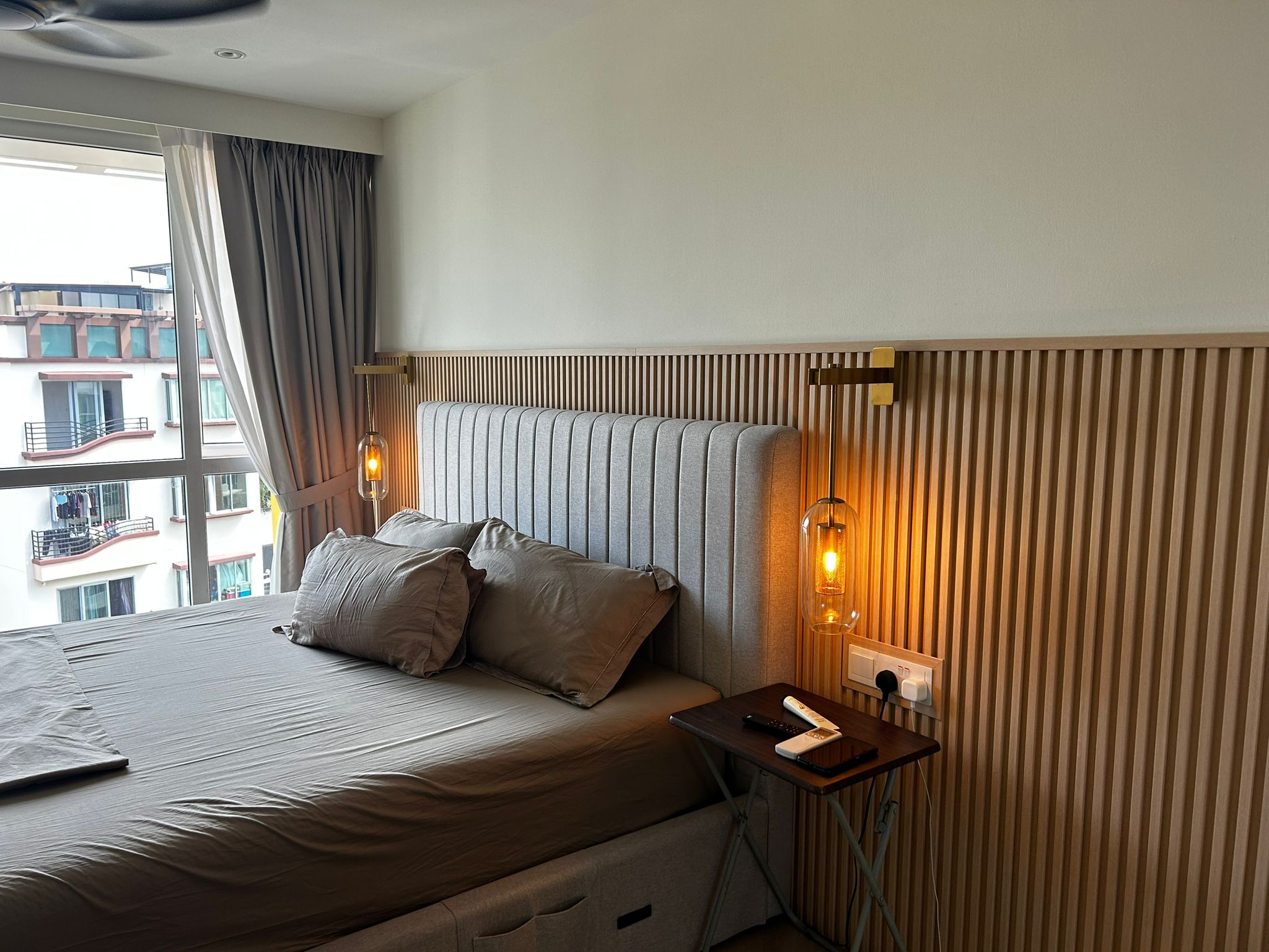 Cozy bedroom with a tufted headboard, warm wall sconces, and a large window with soft gray curtains.