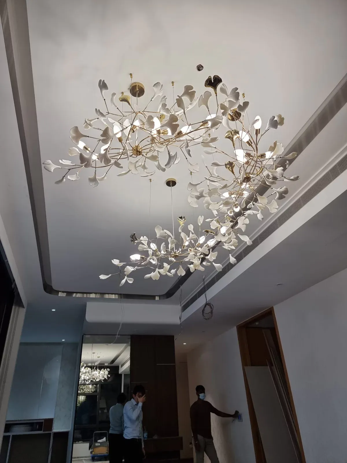 {"default":"Striking ceiling light installation in a modern hallway, featuring an elaborate arrangement of metallic branches and floral elements that spread across the ceiling, illuminated with small lights that cast a warm glow. "}