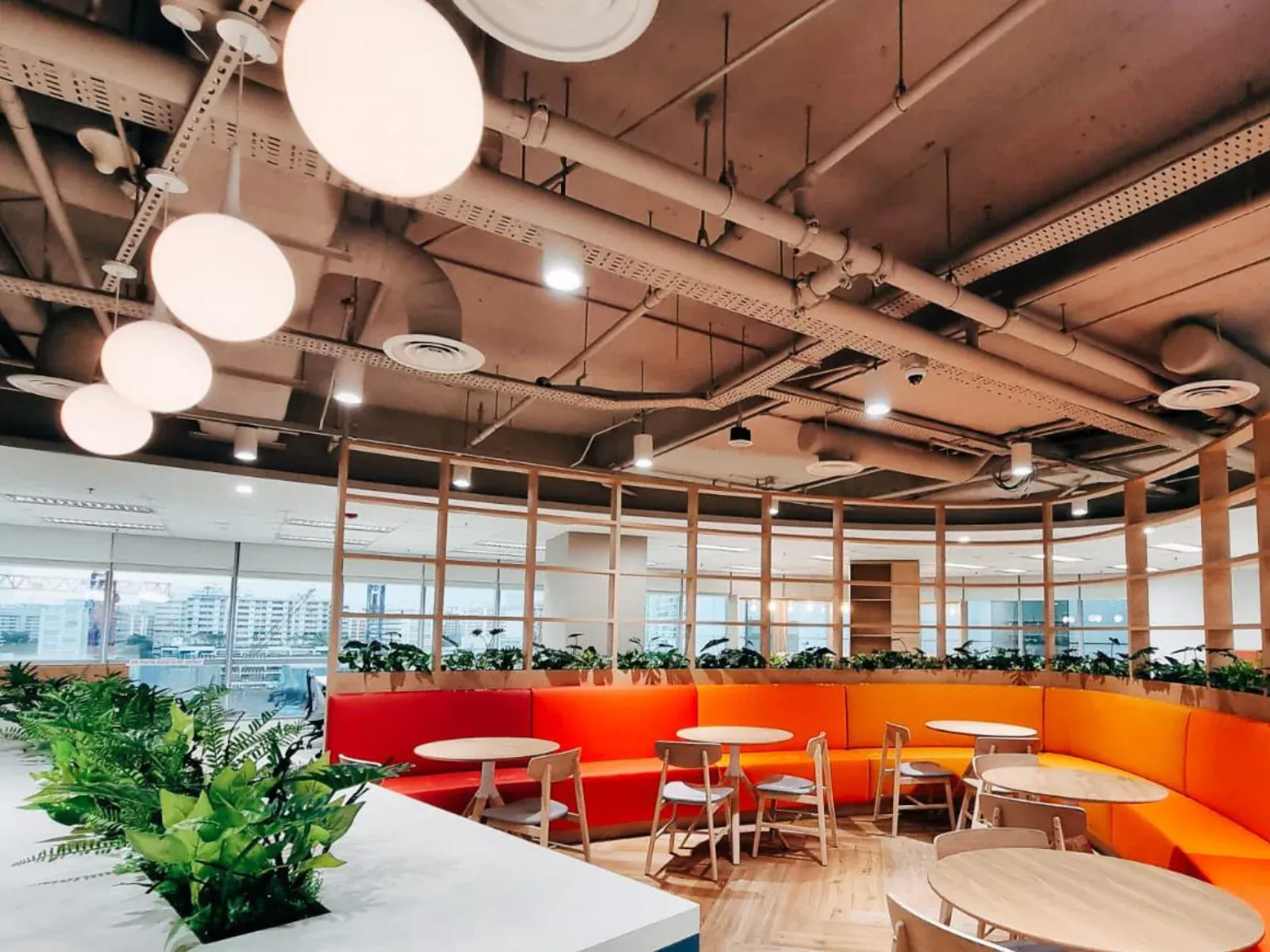 {"default":"Bright and modern cafeteria interior with large globe pendant lights hanging from the ceiling. The space features vibrant orange and white dining tables and chairs, complemented by lush green plants."}
