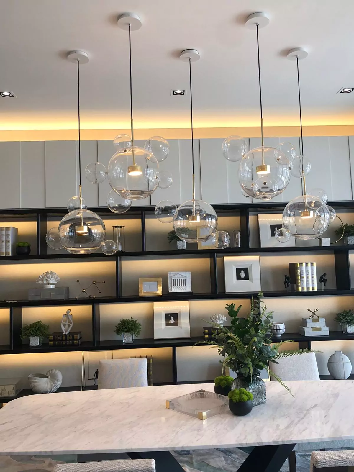 {"default":"Elegant kitchen or dining area featuring a marble countertop with decorative green plants, illuminated by a series of large, clear glass pendant lights hanging from thin, vertical rods. "}