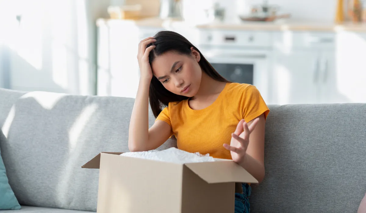 A woman in a yellow shirt looking frustrated while opening a package on her couch.