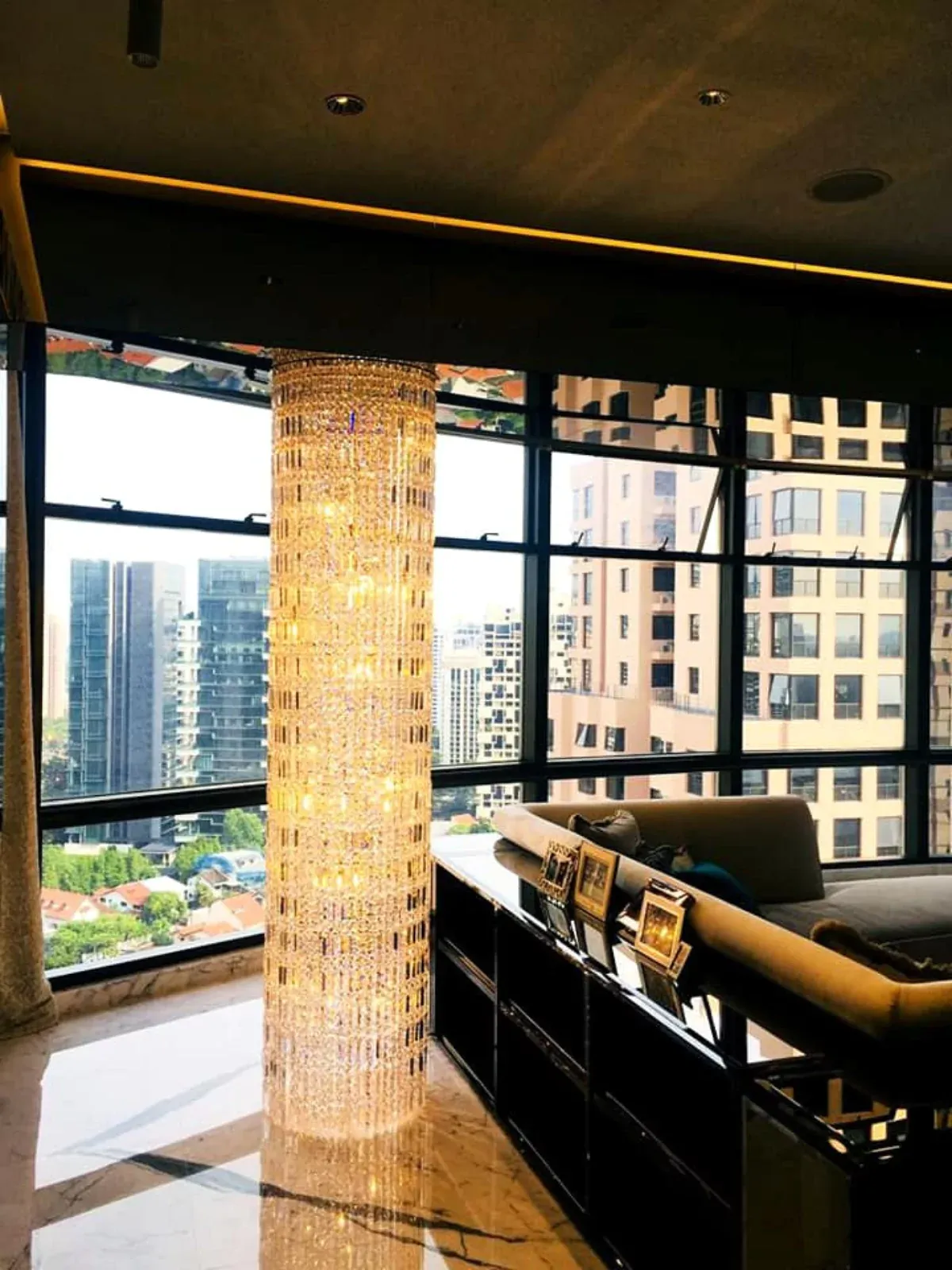 {"default":"Luxurious interior corner featuring a tall, cylindrical chandelier made of shimmering crystals, casting a warm glow next to large windows overlooking an urban skyline. "}