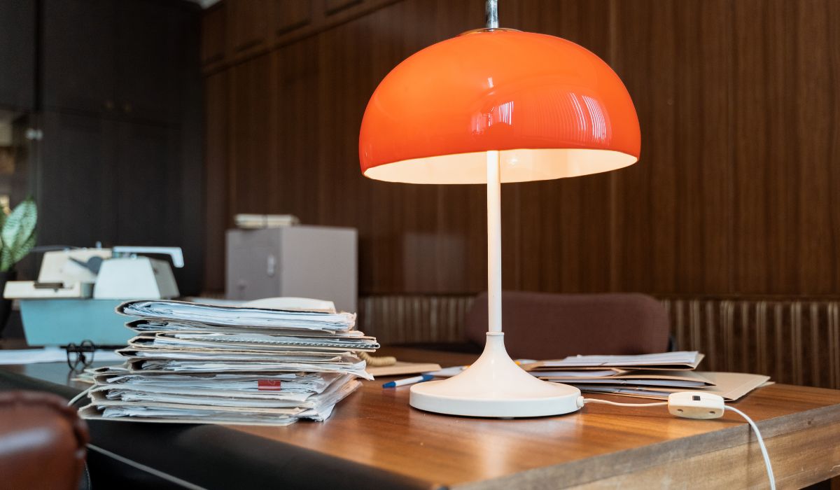 Retro office desk with a red dome lamp, papers, and vintage decor elements.