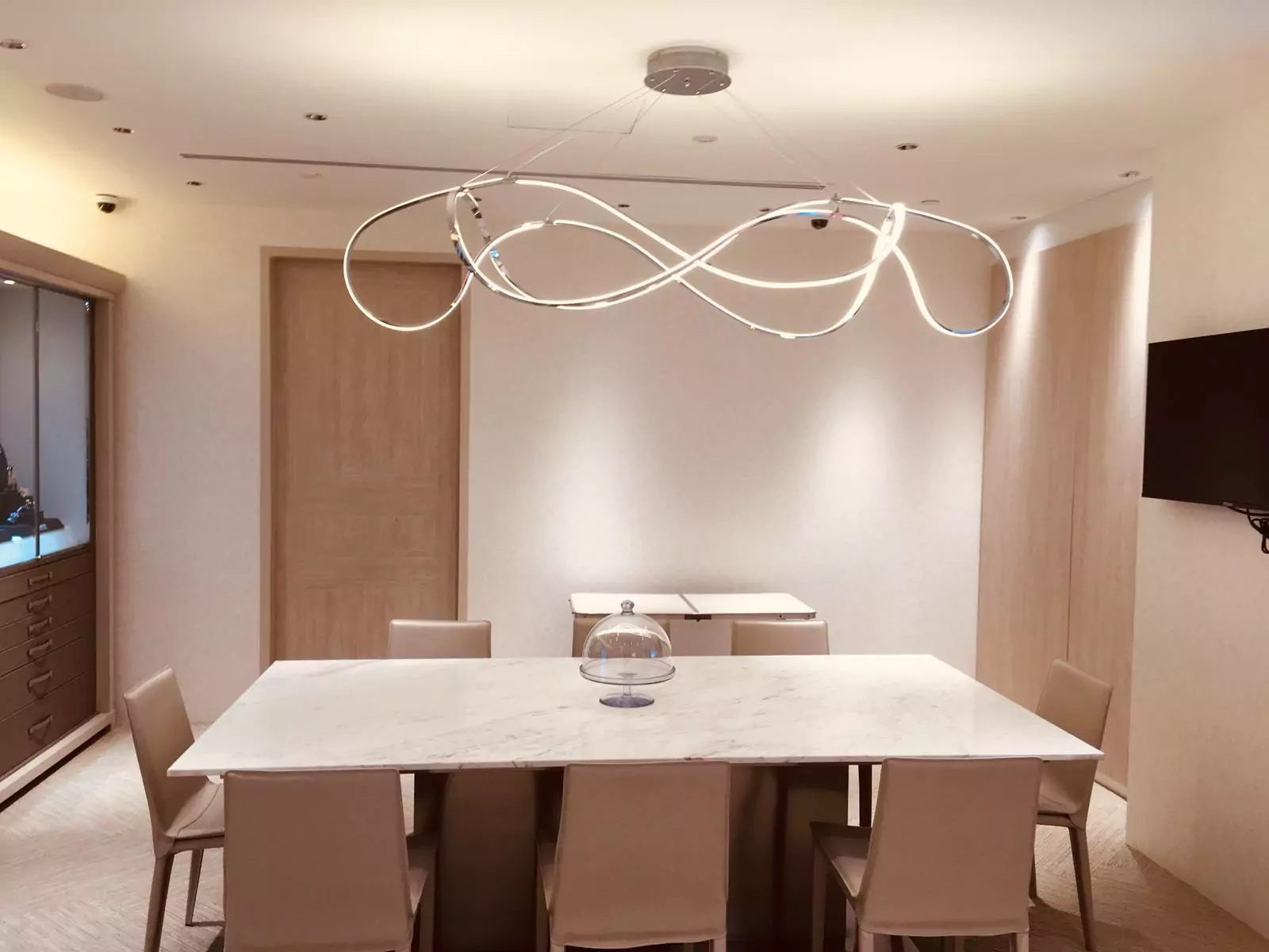 {"default":"Modern dining area featuring a minimalist white table with matching chairs, illuminated by a unique overhead light fixture with intertwined loops, set in a softly lit room with neutral tones."}