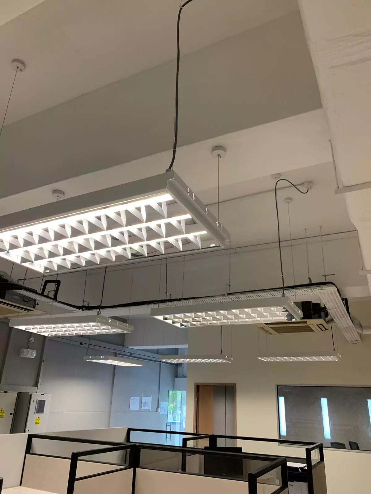 {"default":"Modern office space with an industrial look, featuring a series of rectangular LED light panels suspended from a high ceiling, casting bright light over a minimalist black railing and the office floor below."}