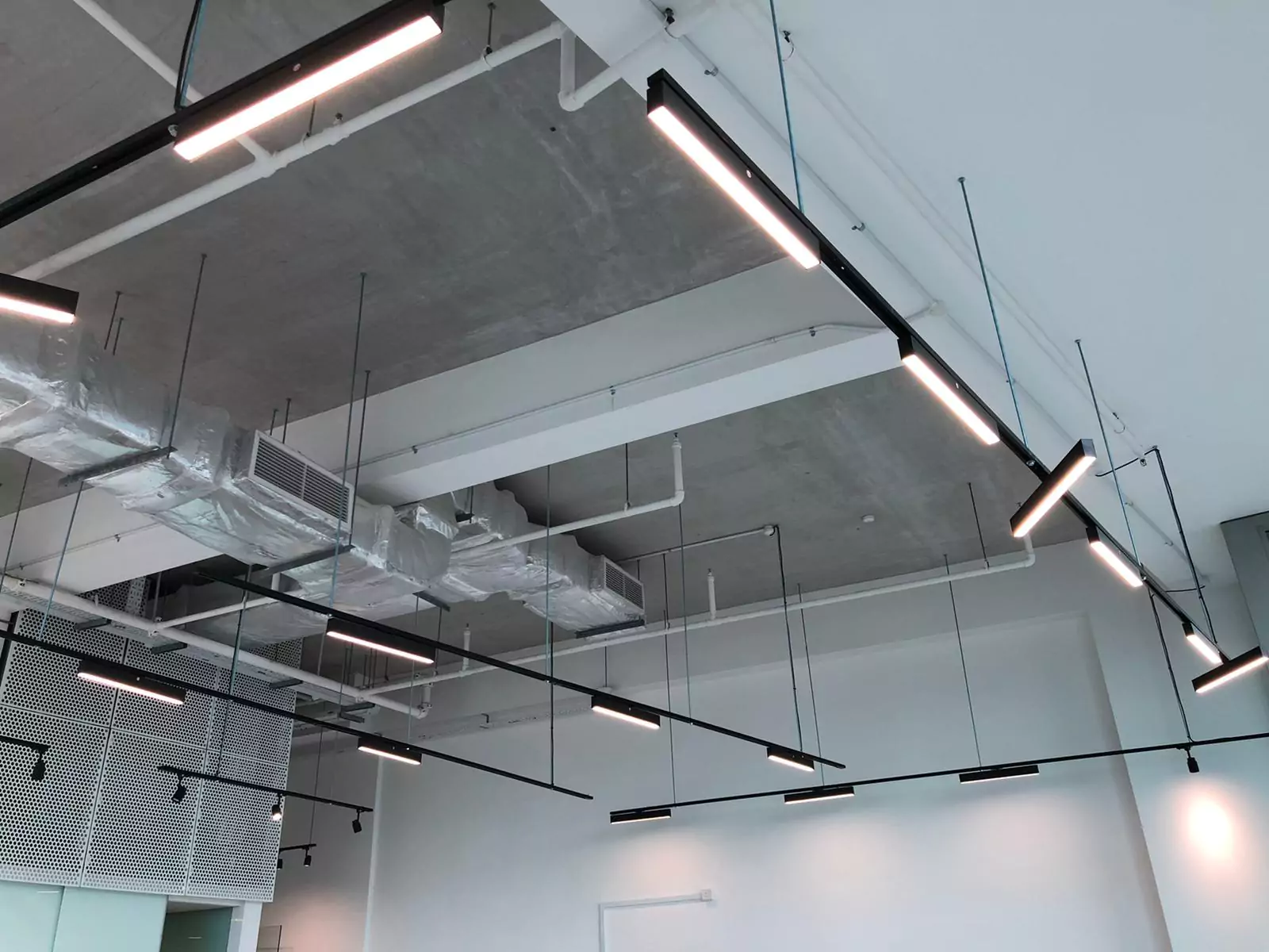 {"default":"Contemporary office space illuminated by suspended track lighting, featuring several linear lights attached to black metal rods that crisscross over an exposed ceiling structure. "}