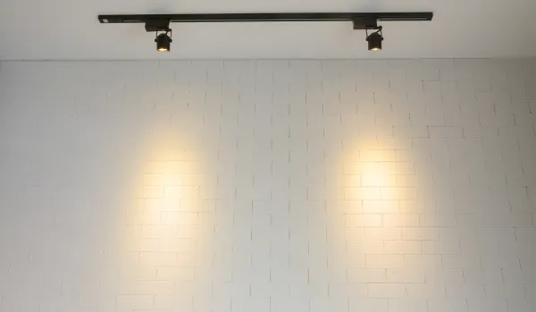 Minimalist black track light fixture with two adjustable spotlights casting warm beams on a white brick wall.