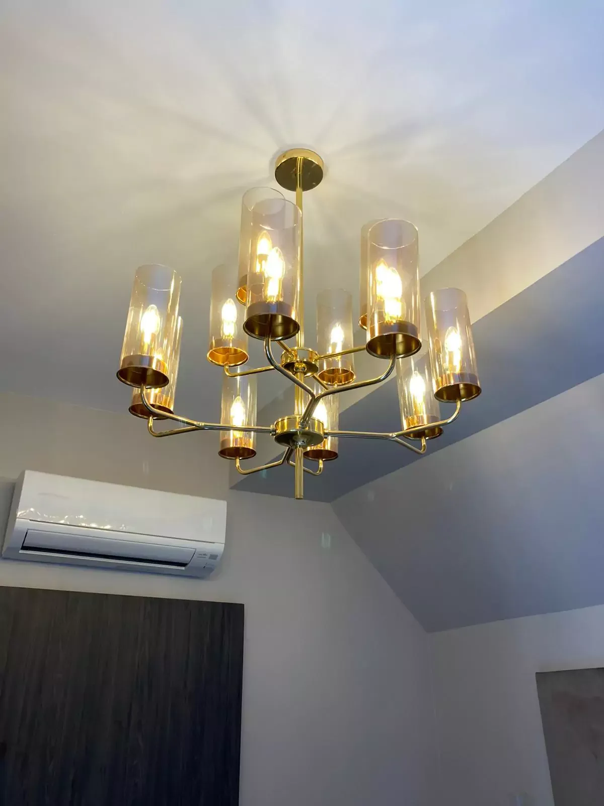 {"default":"Modern chandelier featuring a gold frame with multiple arms, each supporting a tall, cylindrical glass shade. The warm light emitted from the bulbs within the shades casts a soft glow, enhancing the sophisticated ambiance of the room. "}