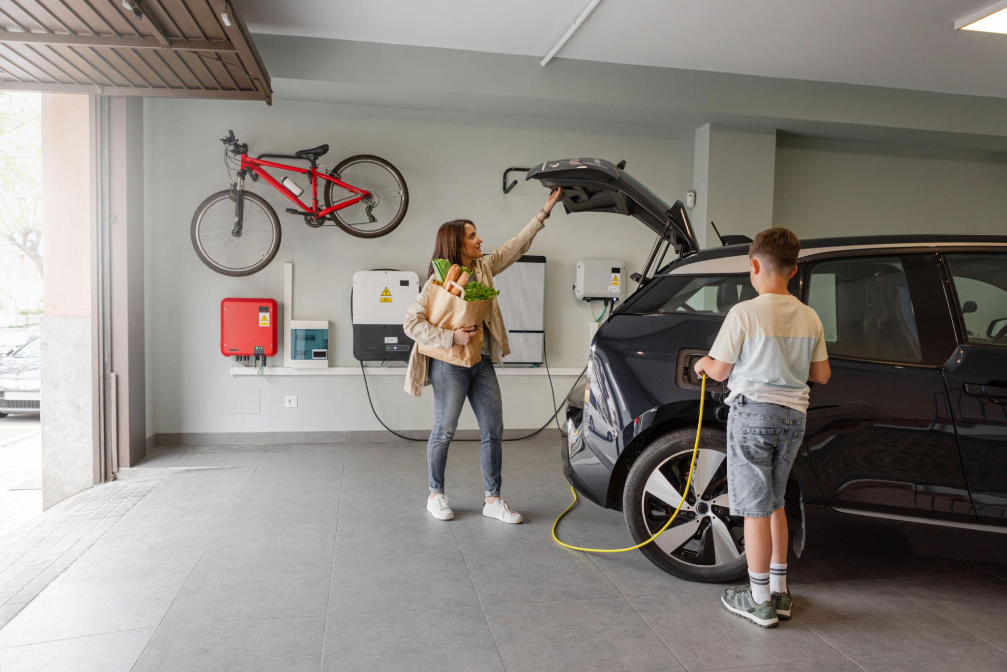 Family in a garage charging an electric car next to wall-mounted solar energy storage units.