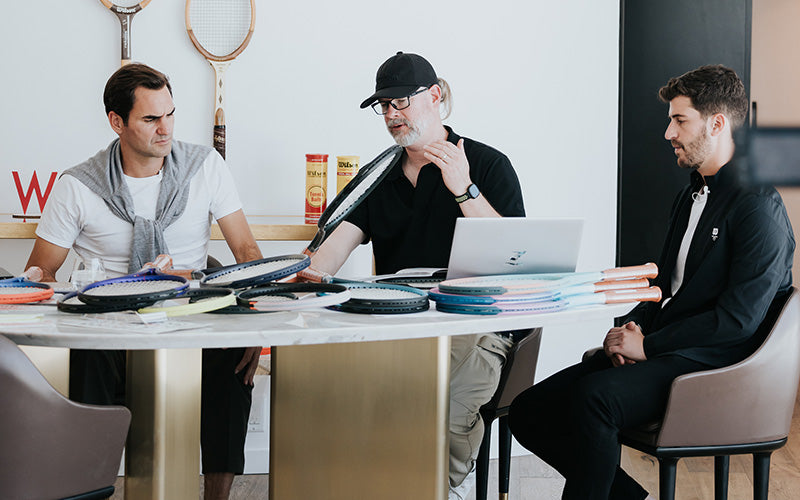Roger Federer and two other men sat around a round table, talking about a racket which one of the men is holding