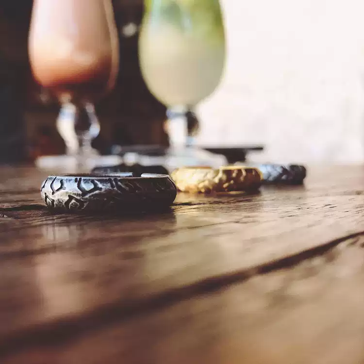 A row of motorcycle tire tread rings in various metallic finishes lined up on a rustic wooden table with blurred drinks in the background.