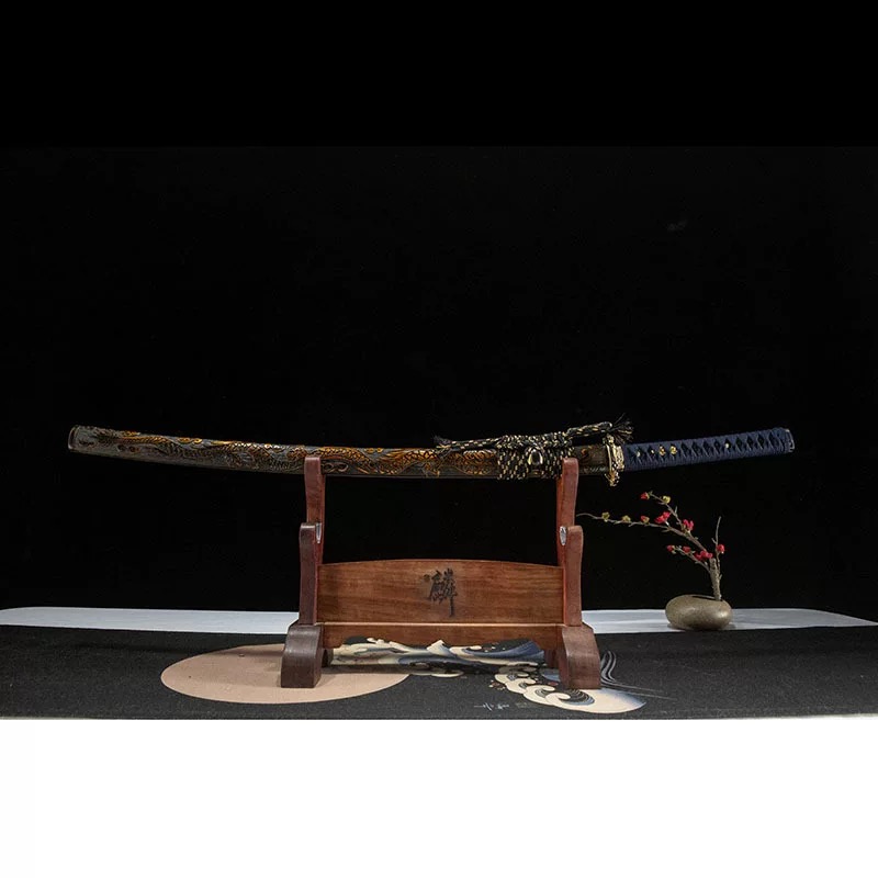 A complete Japanese katana sword in its ornate scabbard, displayed on a traditional wooden sword stand against a black background. Note the blue silk hilt wrap and gold accents.