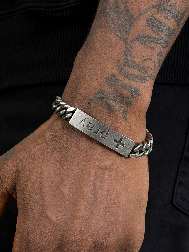 A close-up of a man wearing the SafeHaven Armor Motorcycle Heartbeat chain bracelet featuring a silver metal plate engraved with a cross and the word pray.