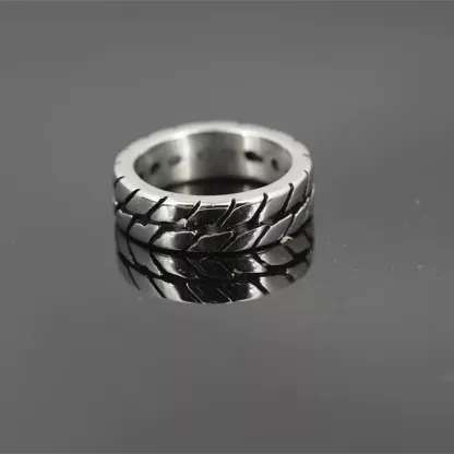 Men's motorcycle tire tread band ring made of durable stainless steel, resting on a dark reflective surface with a clear reflection.