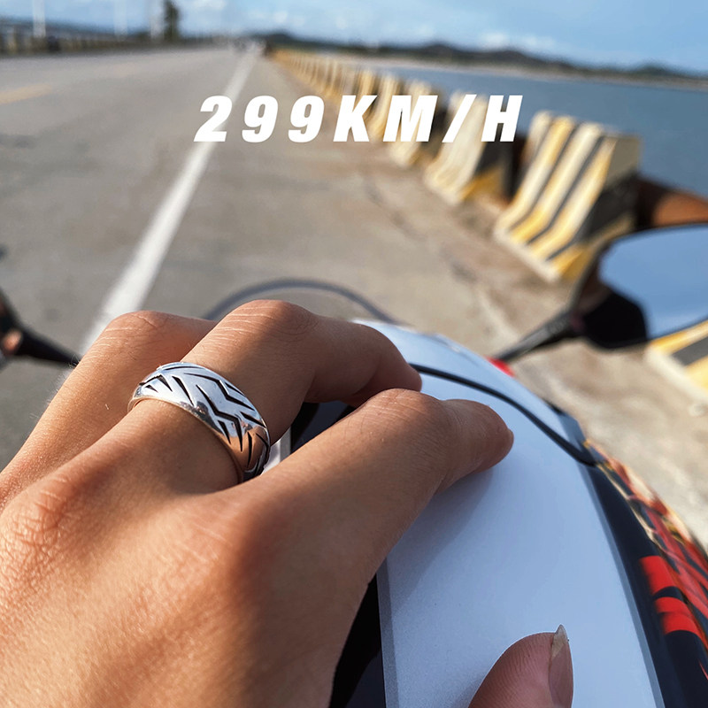 Side view of a silver motorcycle tire tread ring worn on a rider's hand, with a scenic open highway and coastal view in the background.