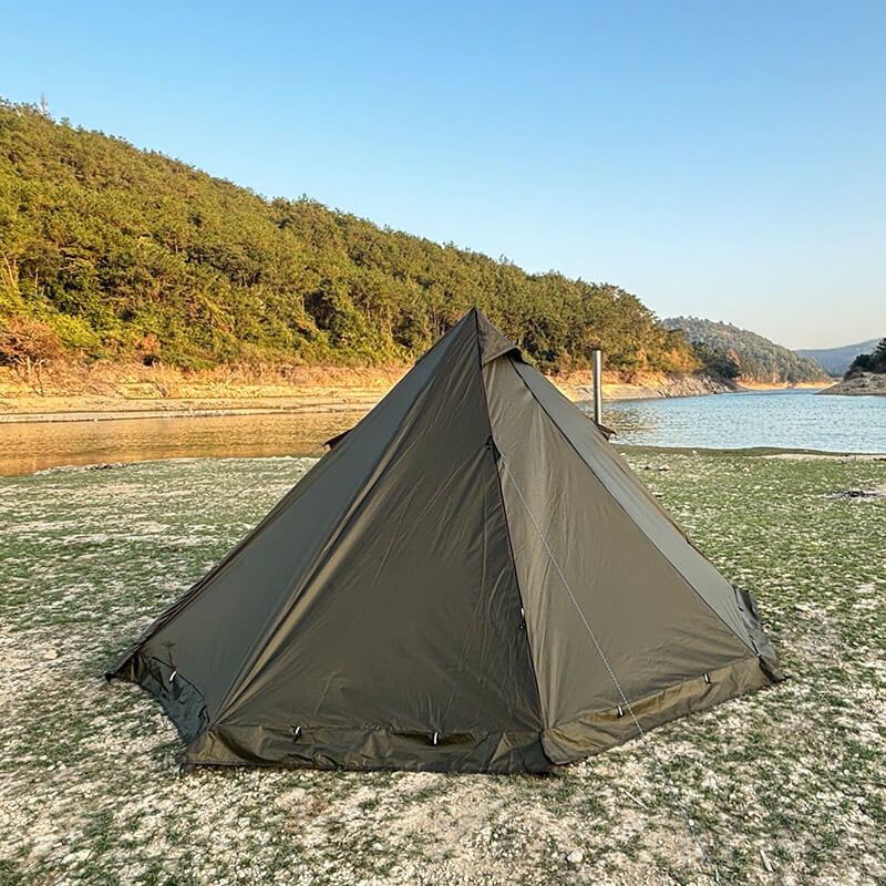 Green pyramid hot tent with a stove jack set up by a lake with a scenic mountain backdrop.