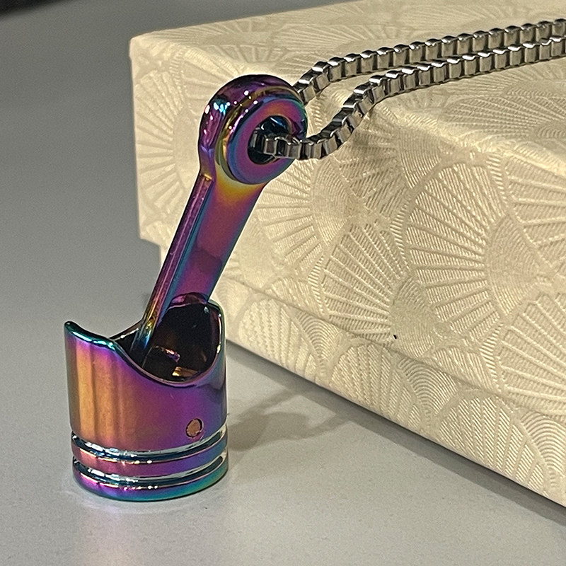 A rainbow iridescent motorcycle piston necklace pendant with a stainless steel box chain resting on a white gift box.
