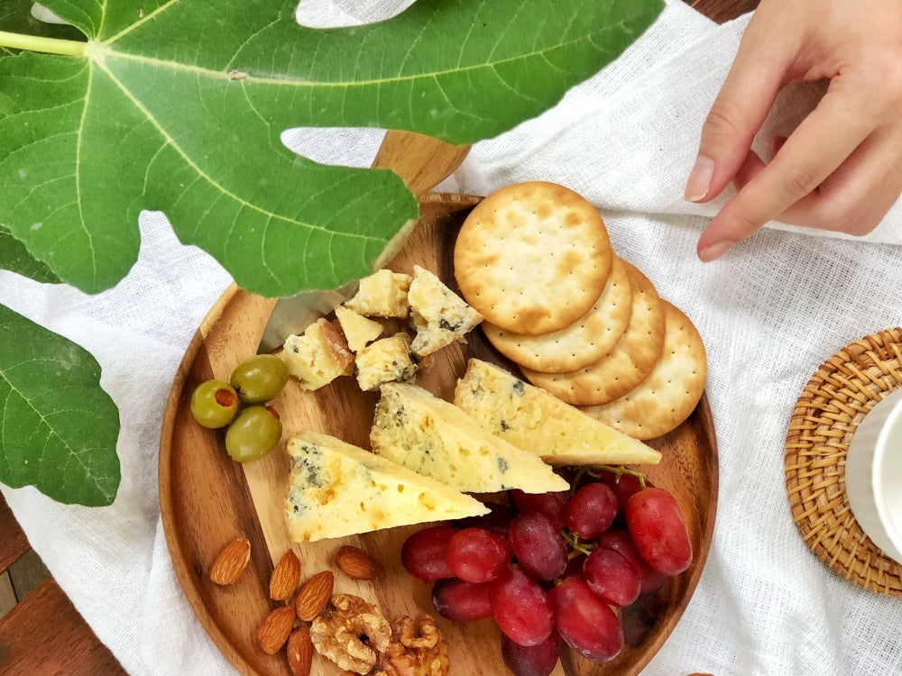 A hand reached out to a small cheese platter with Smoked Melaka cheese slices, grapes, crackers, nuts & olives.