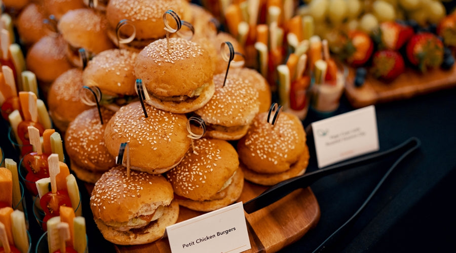 Table spread of petite chicken burgers stacked on wooden board, with a side of small clear cups filled with vegetable sticks
