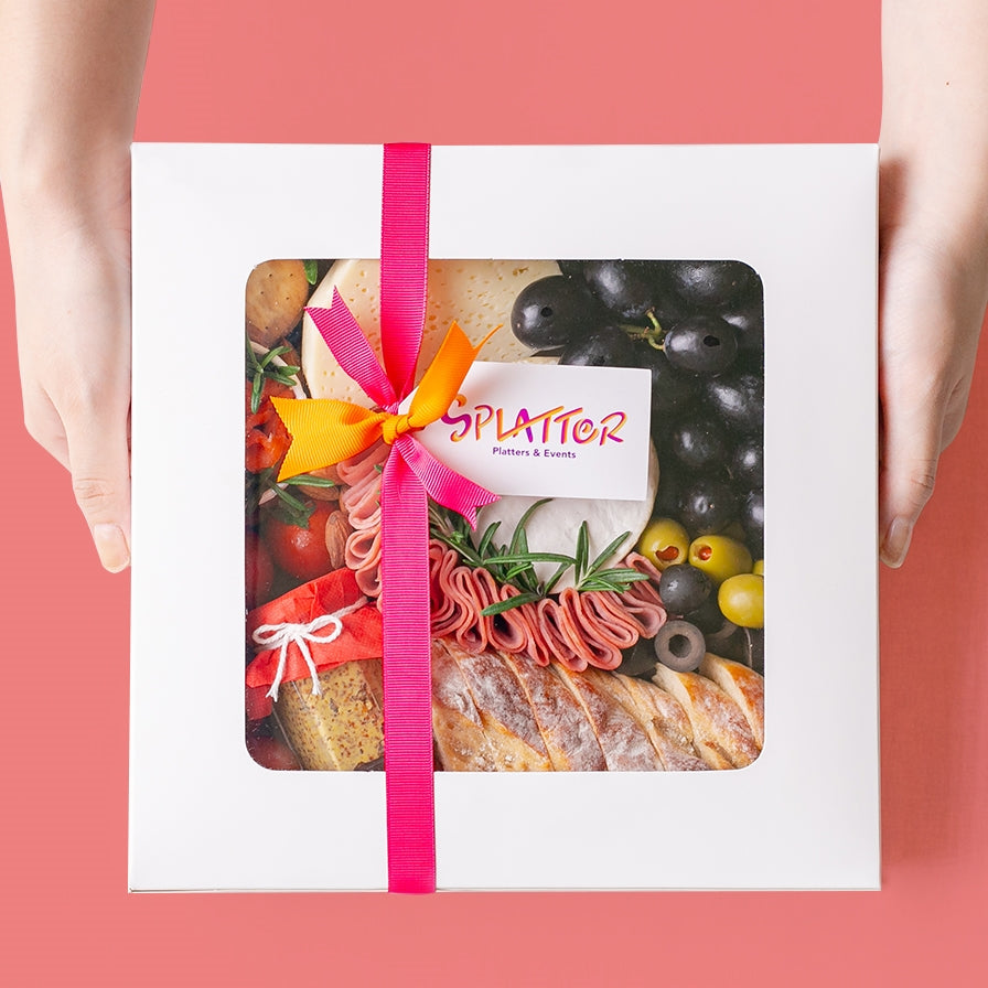 {"default":"Splatter's Cheese & Meat Platter in a covered white box with transparent window that shows most of its content. Pink and orange ribbons form a knot in the middle with a tag branded with Splatter's logo. Two hands hold the side of the box."}

