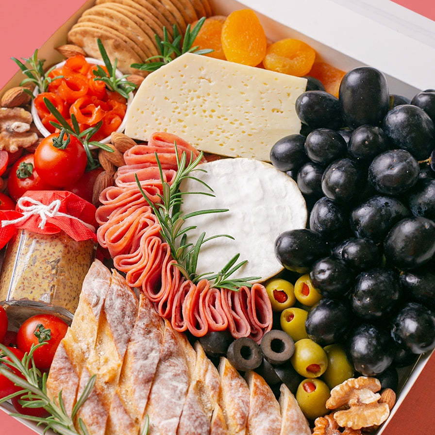 A white square box is packed to the brim with a soft cheese wheel and semi-firm cheese block accompanied by black grapes, cherry tomatoes, green olives, black olives, smoked salmon, turkey rolls, jar of wholegrain mustard, dried apricots, roasted almonds, walnuts, crackers, and baguette slices.