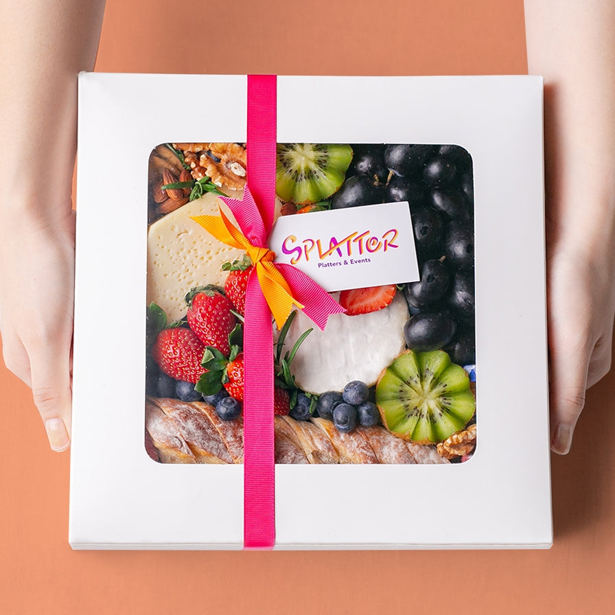 An array of Splatter's Cheese & Fruit Platter ingredients neatly arranged inside a white square box. The box is covered and contents peek through its transparent window. Two hands hold the platter box sides.