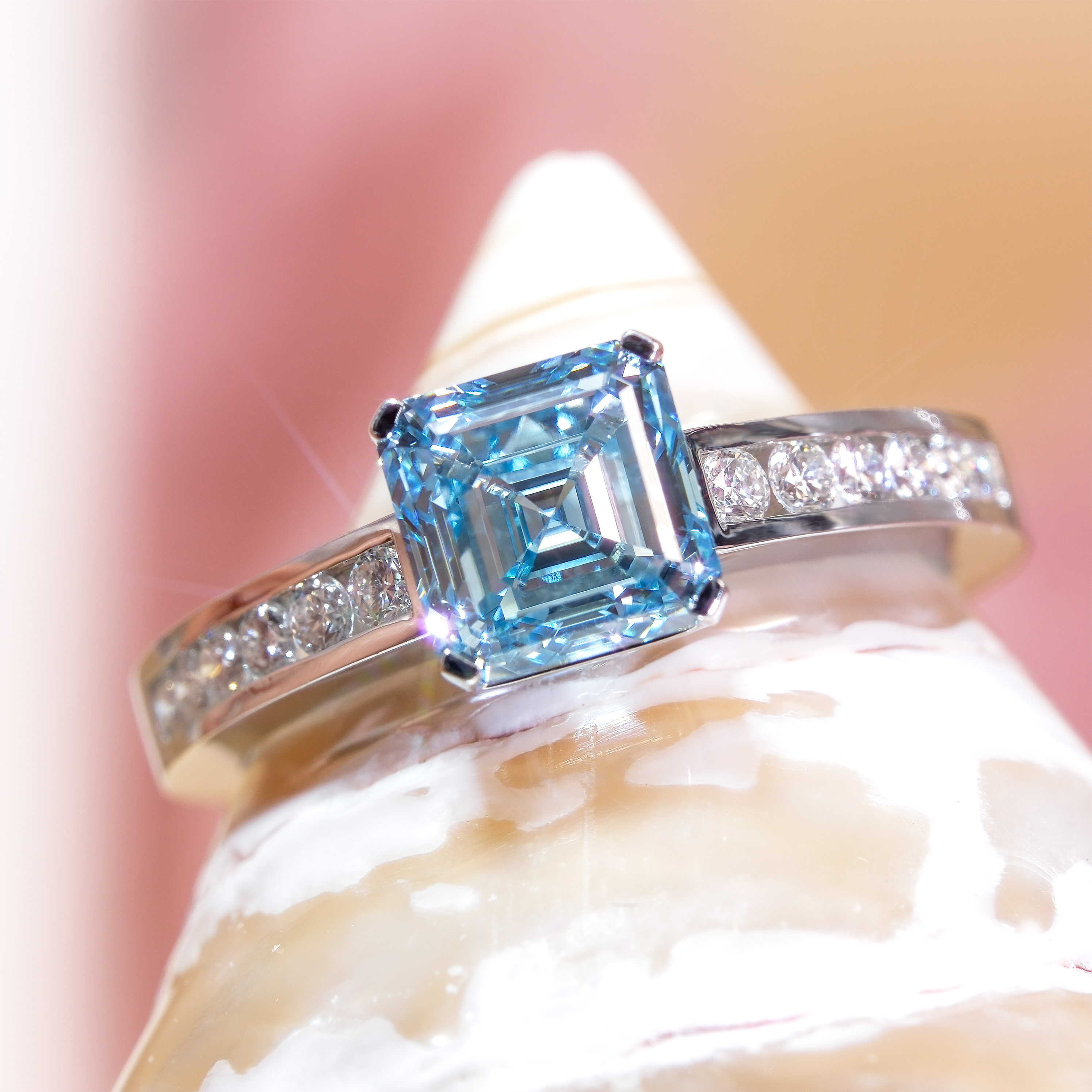 .01 克拉阿斯切形 (Asscher Cut) 培育藍鑽戒指的極致近拍。主石呈現高貴的冰藍色 (Ice Blue)，其獨特的階梯式切割 (Step Cut) 與切角設計，在光線下折射出猶如「萬花筒」般深邃且對稱的光影迷宮。18K 白金戒臂兩側採用槽鑲 (Channel Setting) 工藝，鑲嵌了璀璨的圓形白鑽，與藍色主石形成華麗對比。