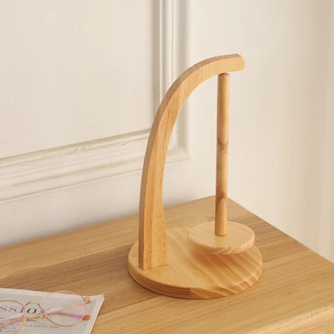 A wooden headphone stand with a curved support and round base sits on a wooden surface beside eyeglasses, a magazine, and the Levitating Premium Yarn Holder for seamless knitting.