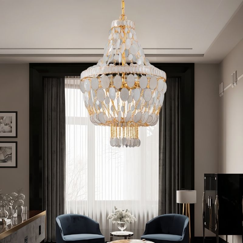 "Tiered chandelier with cascading natural shell elements and gold frame, hanging in a living room with two blue chairs."