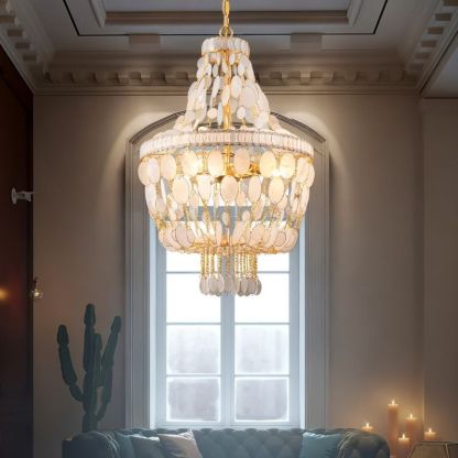"Tiered chandelier with cascading natural shell elements and gold frame, hanging in a living room with a large window."