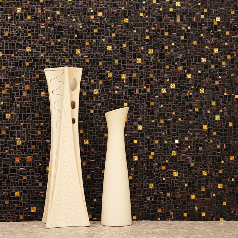 {"default":"黒と金の小さなタイルで構成された壁を背景に、装飾模様のある背の高い白い花瓶と、細身の白い花瓶が石の台の上に並んでいる"}
