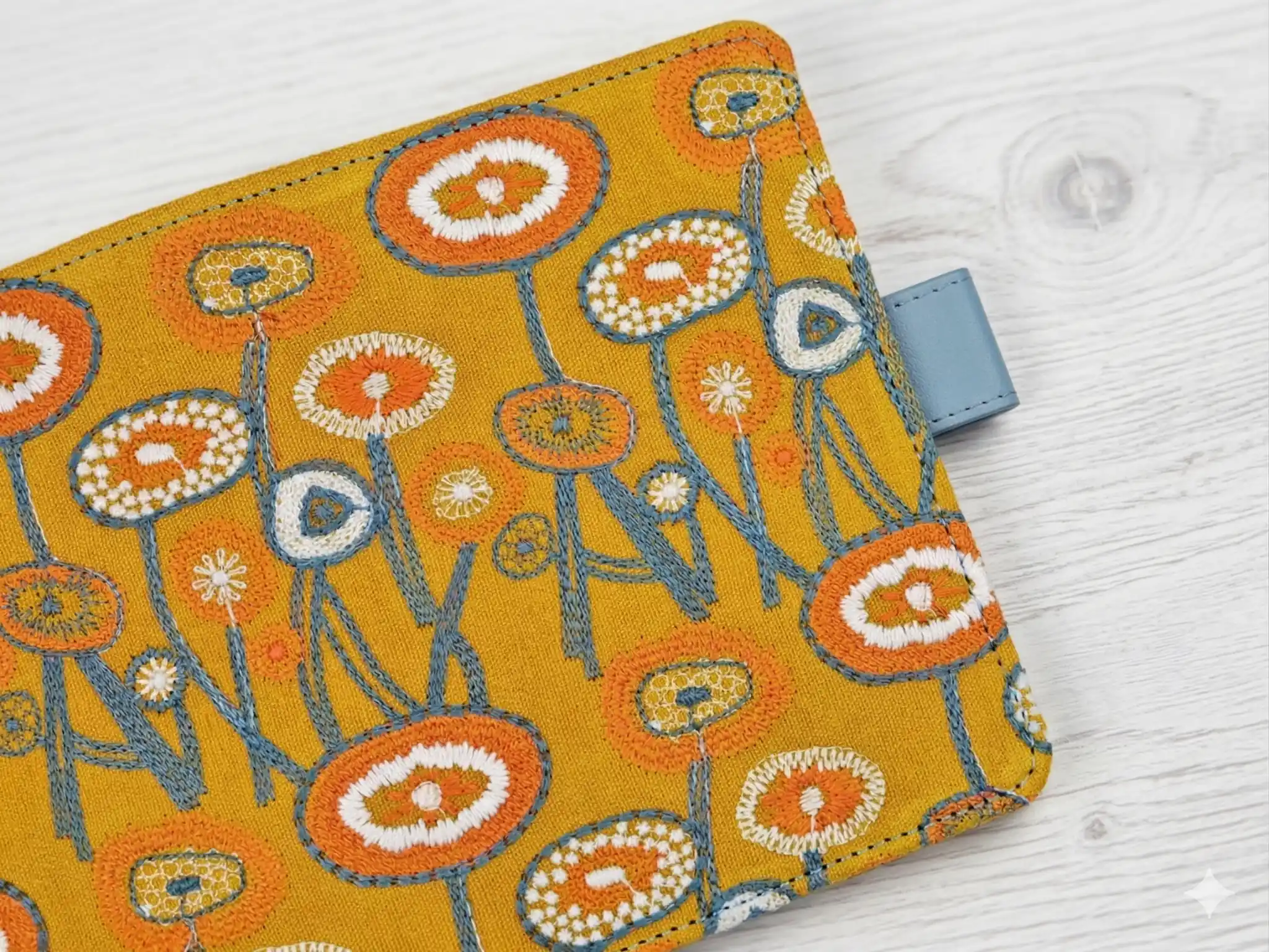 Macro shot of the textured floral embroidery in orange and white threads on yellow fabric, highlighting the tactile handmade quality.