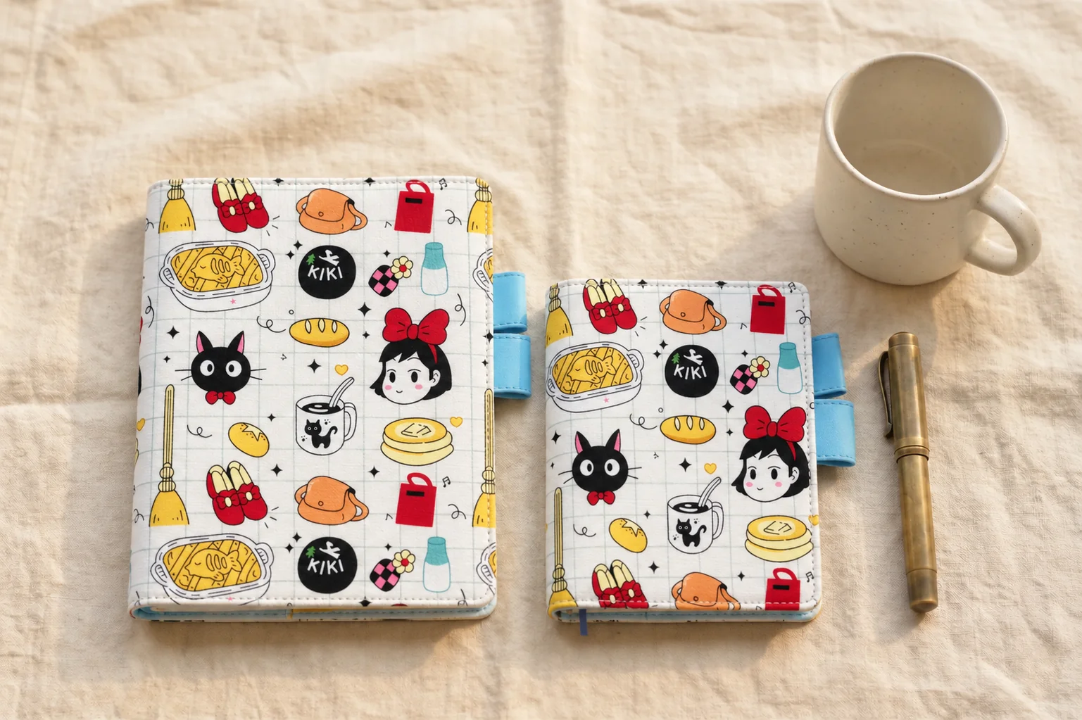 Two black cat and broomstick print planner covers with blue pen loops, arranged on linen beside a mug and brass pen.