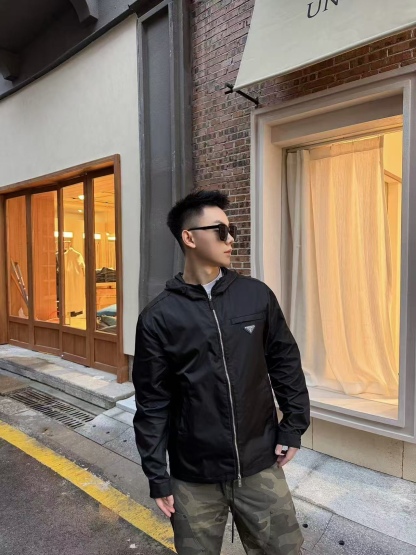 🔝PRADA/普拉達PRD 拉鍊口袋尼龍連帽外套
選用品牌標誌性的再生尼龍面料，與傳統尼龍性能相當，具備高強度，抗撕裂，耐磨損等特性。其質地輕盈，同時又具備防水功能適合多場景需求。抗皺，快乾的特點也易於日常打理維護。
定制三角徽標通過4個0.8mm金屬爪釘固定於衣身，爪釘背面經手工打磨至圓潤，避免刮蹭。下擺彈力抽繩選用7芯高彈尼龍繩，直徑3mm，配合橡膠扣，可實現±5cm長度調節。抽繩孔採用「金屬雞眼+防磨環」雙重保護，雞眼內徑5mm，邊緣光滑無毛刺，反復抽拉500次無變形。
袖口採用紐扣調