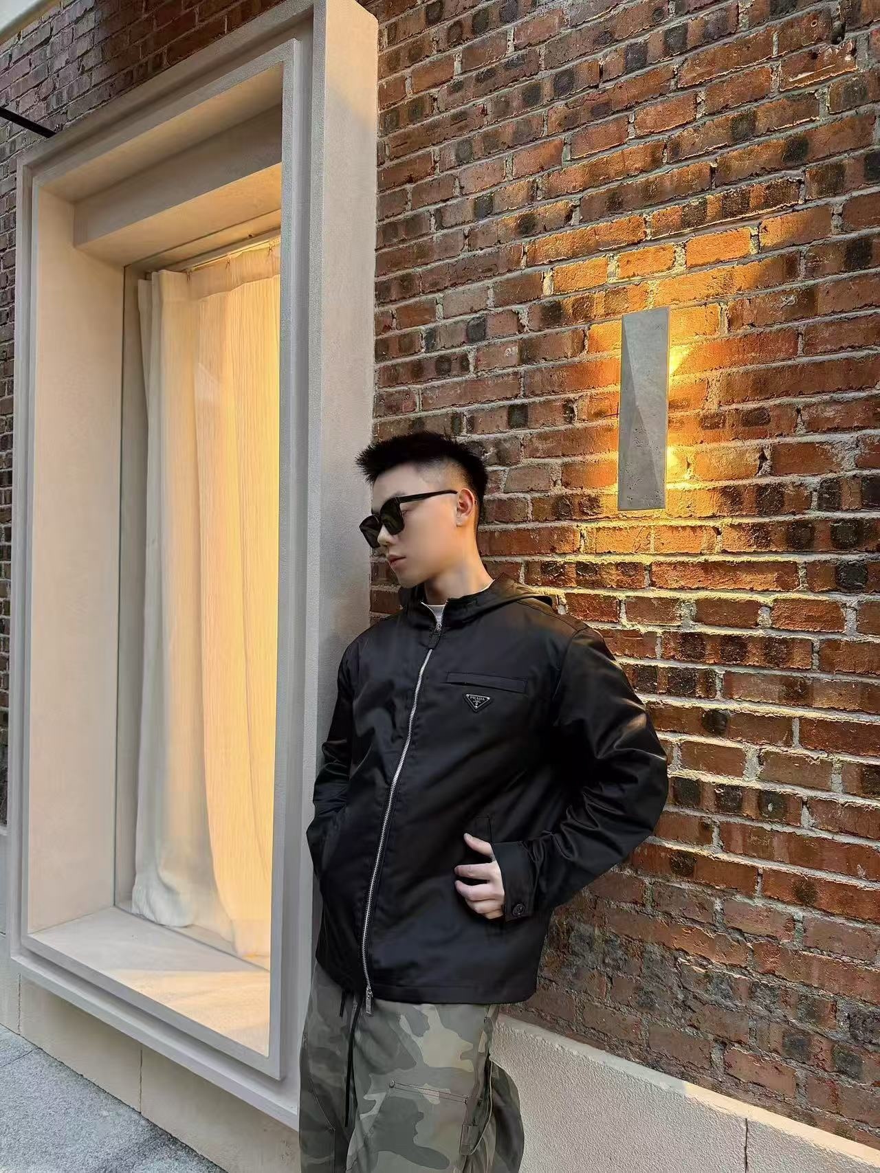 🔝PRADA/普拉達PRD 拉鍊口袋尼龍連帽外套
選用品牌標誌性的再生尼龍面料，與傳統尼龍性能相當，具備高強度，抗撕裂，耐磨損等特性。其質地輕盈，同時又具備防水功能適合多場景需求。抗皺，快乾的特點也易於日常打理維護。
定制三角徽標通過4個0.8mm金屬爪釘固定於衣身，爪釘背面經手工打磨至圓潤，避免刮蹭。下擺彈力抽繩選用7芯高彈尼龍繩，直徑3mm，配合橡膠扣，可實現±5cm長度調節。抽繩孔採用「金屬雞眼+防磨環」雙重保護，雞眼內徑5mm，邊緣光滑無毛刺，反復抽拉500次無變形。
袖口採用紐扣調