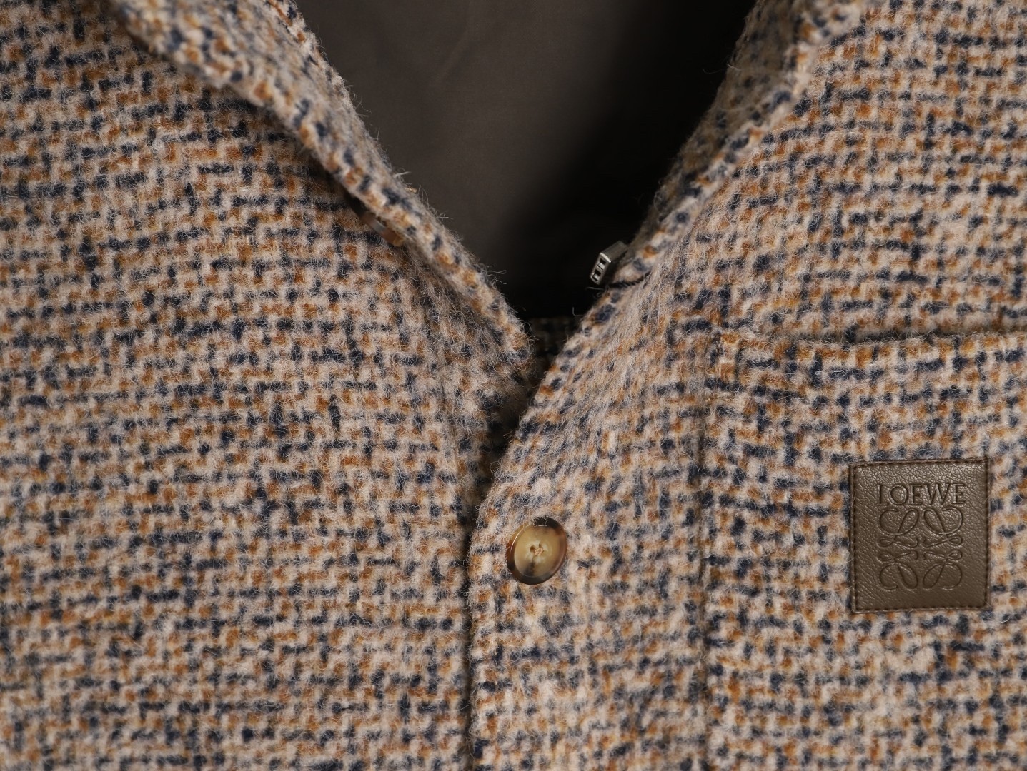 🔝LOEWE羅意威 皮章口袋羊毛混紡外套
採用精心配比的混紡面料，羊毛提供天然保暖與挺括廓形，馬海毛賦予面料細膩蓬松的絨毛感，少量錦綸的餵入則增強耐磨性與抗皺性，三者融合打造中克重的理想厚度，兼顧溫度與輕盈。通過“雙面磨毛”工藝處理，面料表面呈現豐盈的短絨毛肌理，觸感如羊絨般柔軟親膚，同時保留羊毛混紡的筋骨感。微型格紋圖案以沙米色為基底，交織灰、棕、藍三色紗線，通過色織工藝織就，微型格紋尺寸統一，細膩而不張揚，呈現低調的復古韻味。
面料經過3道磨毛工序，確保絨毛長度均勻（约0.3mm）