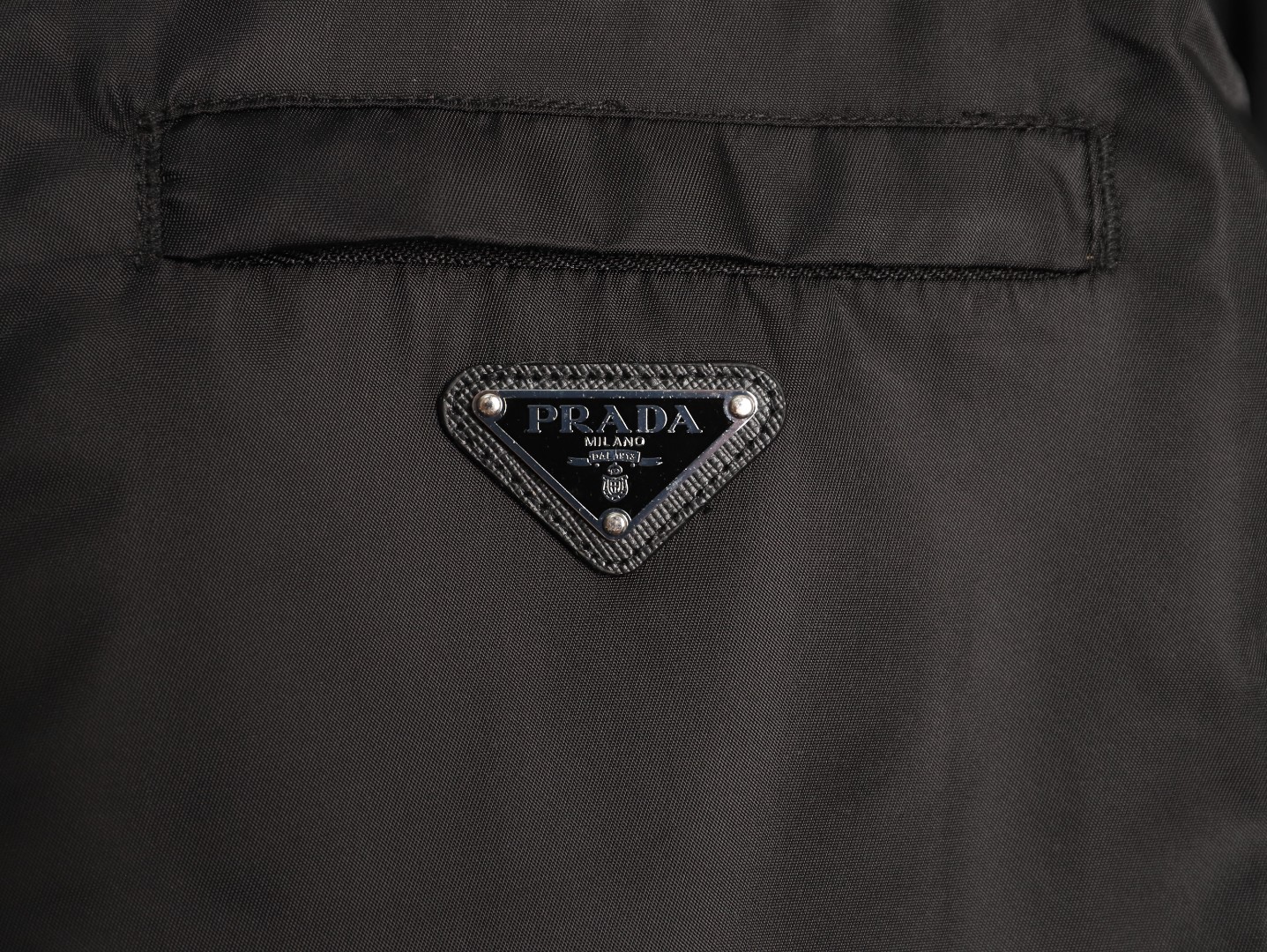 🔝PRADA/普拉達PRD 拉鍊口袋尼龍連帽外套
選用品牌標誌性的再生尼龍面料，與傳統尼龍性能相當，具備高強度，抗撕裂，耐磨損等特性。其質地輕盈，同時又具備防水功能適合多場景需求。抗皺，快乾的特點也易於日常打理維護。
定制三角徽標通過4個0.8mm金屬爪釘固定於衣身，爪釘背面經手工打磨至圓潤，避免刮蹭。下擺彈力抽繩選用7芯高彈尼龍繩，直徑3mm，配合橡膠扣，可實現±5cm長度調節。抽繩孔採用「金屬雞眼+防磨環」雙重保護，雞眼內徑5mm，邊緣光滑無毛刺，反復抽拉500次無變形。
袖口採用紐扣調