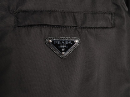 🔝PRADA/普拉達PRD 拉鍊口袋尼龍連帽外套
選用品牌標誌性的再生尼龍面料，與傳統尼龍性能相當，具備高強度，抗撕裂，耐磨損等特性。其質地輕盈，同時又具備防水功能適合多場景需求。抗皺，快乾的特點也易於日常打理維護。
定制三角徽標通過4個0.8mm金屬爪釘固定於衣身，爪釘背面經手工打磨至圓潤，避免刮蹭。下擺彈力抽繩選用7芯高彈尼龍繩，直徑3mm，配合橡膠扣，可實現±5cm長度調節。抽繩孔採用「金屬雞眼+防磨環」雙重保護，雞眼內徑5mm，邊緣光滑無毛刺，反復抽拉500次無變形。
袖口採用紐扣調