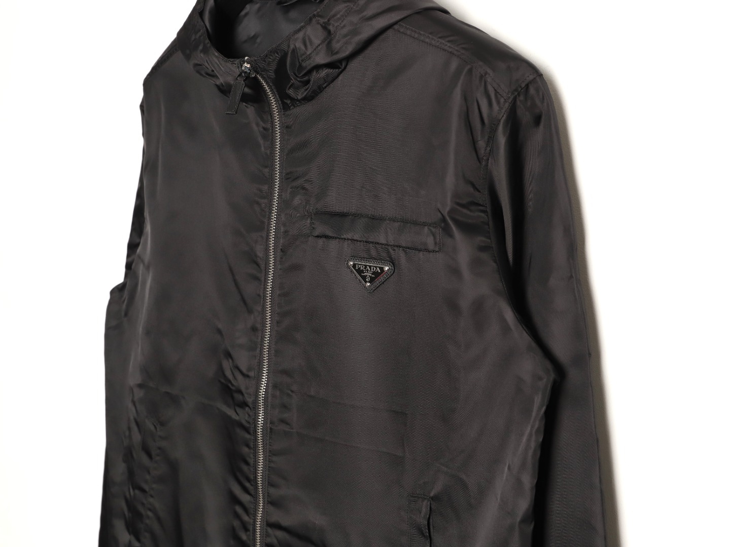 🔝PRADA/普拉達PRD 拉鍊口袋尼龍連帽外套
選用品牌標誌性的再生尼龍面料，與傳統尼龍性能相當，具備高強度，抗撕裂，耐磨損等特性。其質地輕盈，同時又具備防水功能適合多場景需求。抗皺，快乾的特點也易於日常打理維護。
定制三角徽標通過4個0.8mm金屬爪釘固定於衣身，爪釘背面經手工打磨至圓潤，避免刮蹭。下擺彈力抽繩選用7芯高彈尼龍繩，直徑3mm，配合橡膠扣，可實現±5cm長度調節。抽繩孔採用「金屬雞眼+防磨環」雙重保護，雞眼內徑5mm，邊緣光滑無毛刺，反復抽拉500次無變形。
袖口採用紐扣調